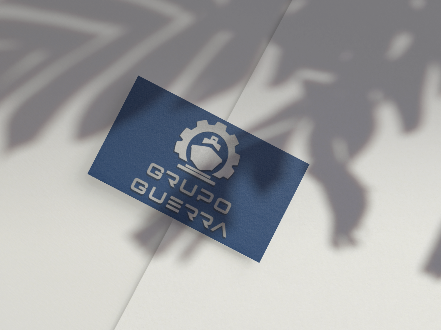 chão branco com o cartão de visita como o logotipo do Grupo Guerra, composto por um ícone circular com uma engrenagem e um navio estilizado no centro, acompanhado do nome “GRUPO GUERRA” em letras maiúsculas brancas sobre um fundo azul.